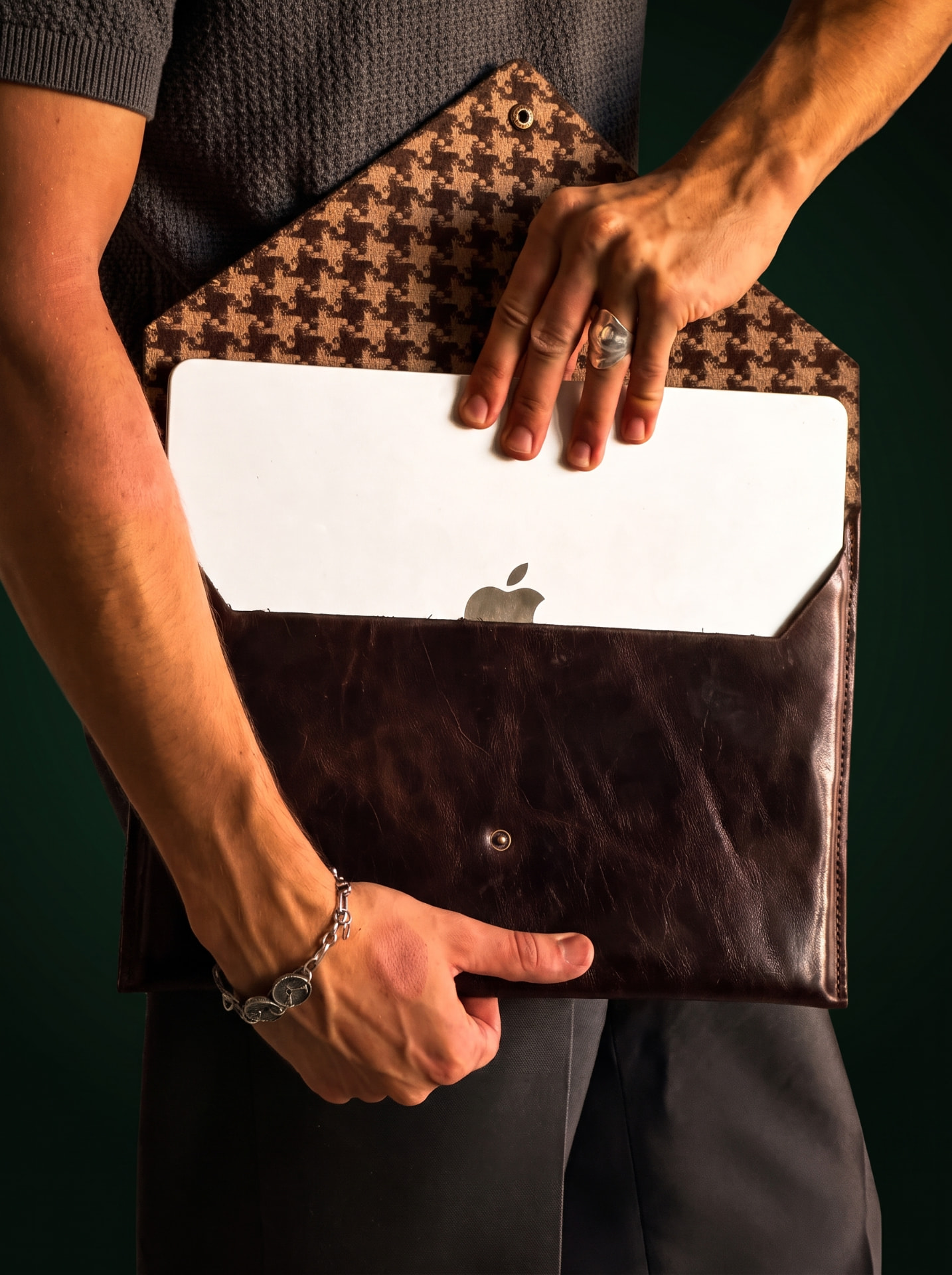 Man in a grey knit polo holding a dark brown leather MacLeather envelope with a MacBook inside, houndstooth lining and green backdrop