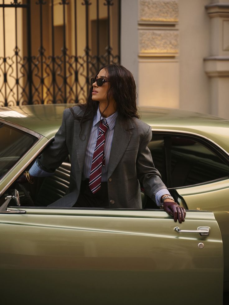 Woman in a tailored grey suit and striped tie with sunglasses, stepping out of a vintage sage-green car